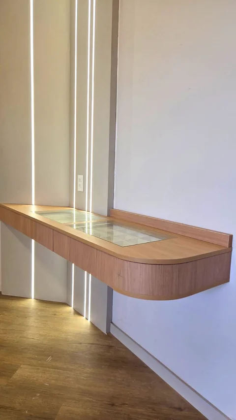 Aparador amadeirado com bordas curvas, tampo de vidro e iluminação vertical na parede.