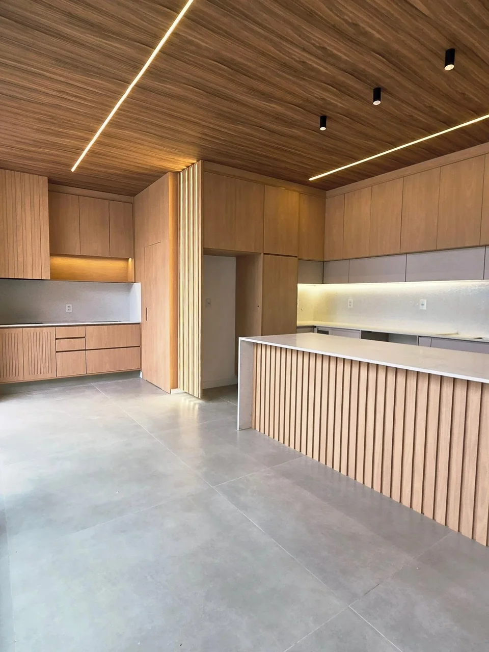 Cozinha Arko com ilha e painéis ripados, armários amadeirados e iluminação integrada ao forro.